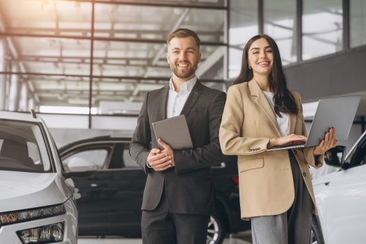 Super sales team in dealership, two consultants or managers in elegant suits with laptop and tablet in arms in car dealership